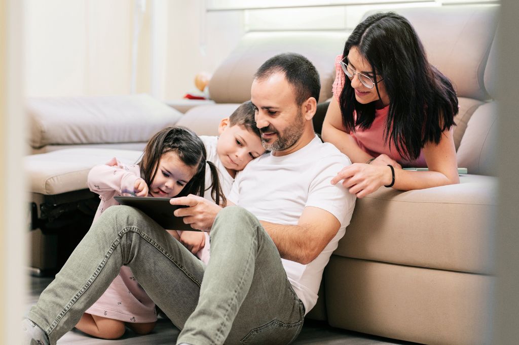 Man sat leaning against a couch watching a tablet with his children and female partner