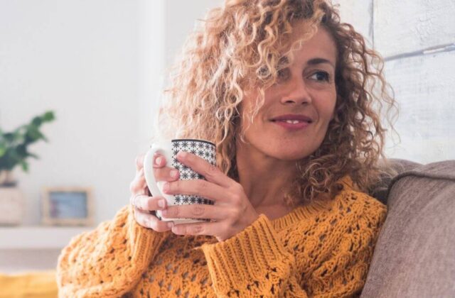 Woman with curly hair and mustard jumper sat drinking a hot drink from a mug