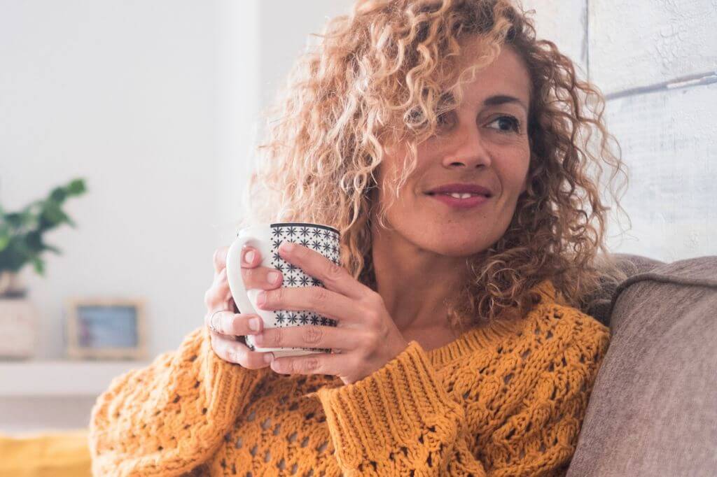 Woman with curly hair and mustard jumper sat drinking a hot drink from a mug